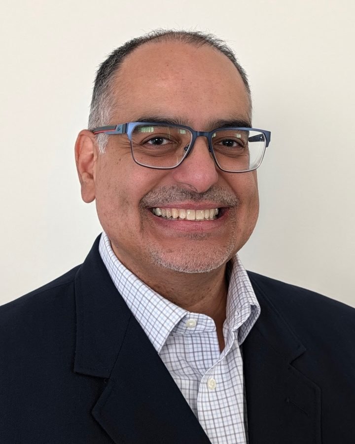 Pedro M. Morales, smiling, wearing blue-framed glasses, a navy blazer, and an open-collar check shirt.